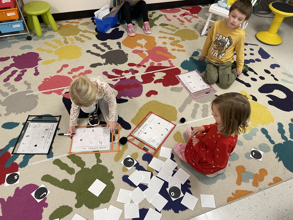 Four young children on a colorful, patterned rug in a classroom, engaged in a literacy activity. Two children are writing on clear sheet protectors with letter tracing worksheets, and two are looking at cards and blank squares on the floor. 