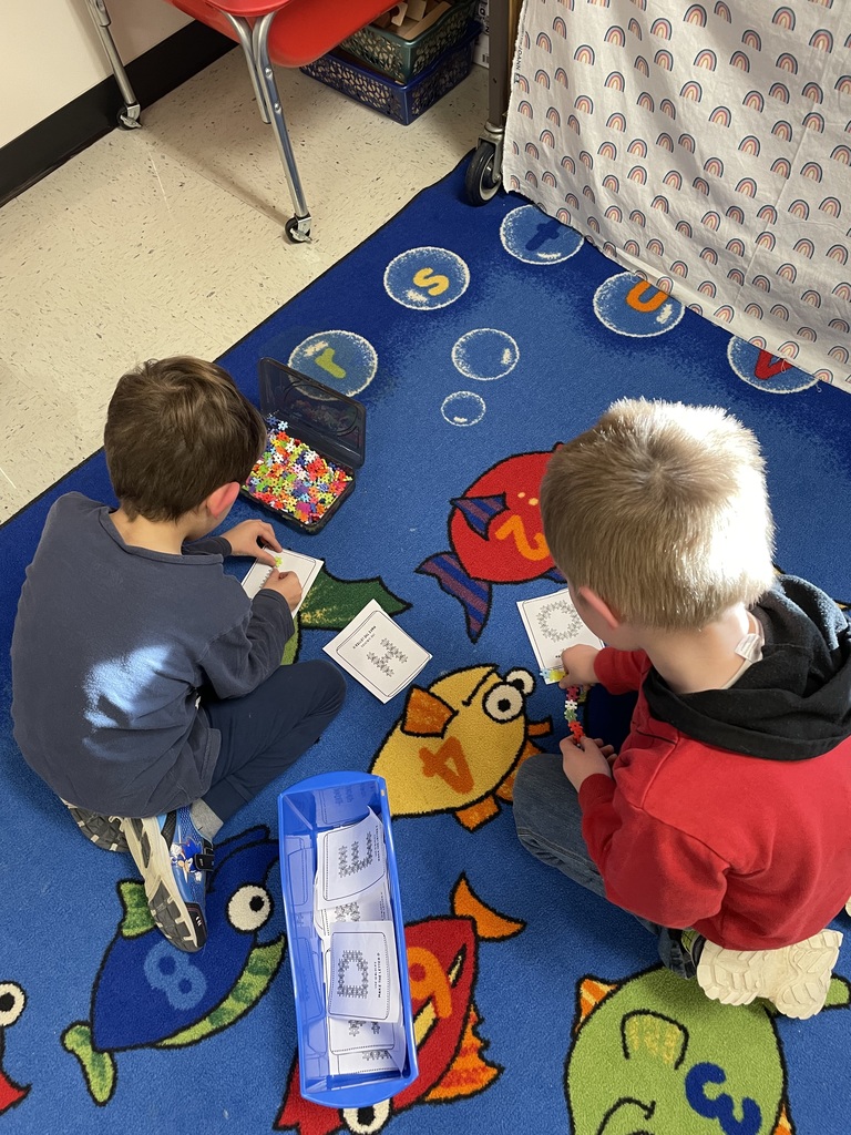  Two young boys sitting on a blue rug with an underwater fish theme, focused on a fine motor skill or math activity. They are using small, colorful plastic connecting pieces from an open bin to complete tasks on printed cards.  