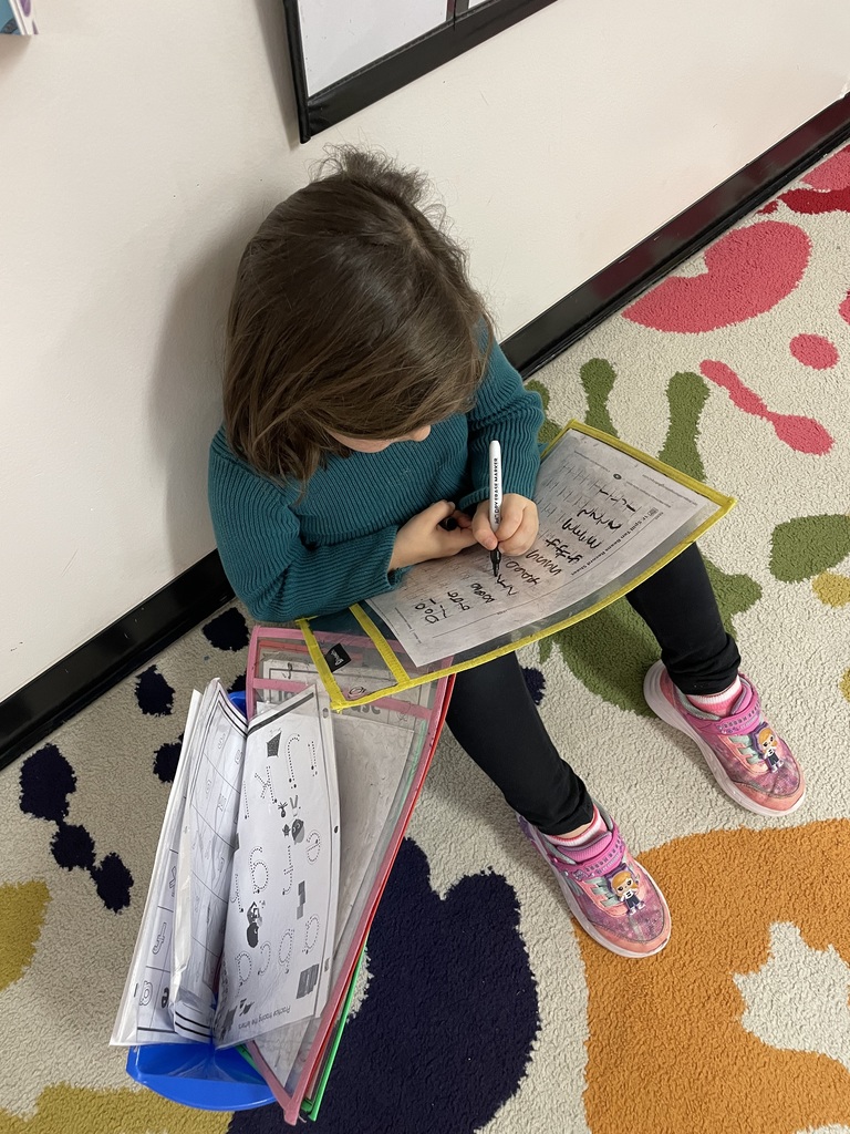 A young girl with dark hair, wearing a green top and black leggings, sitting on a colorful rug while practicing writing. She is intently tracing letters or numbers on a worksheet secured in a clear yellow sleeve, with a stack of similar worksheets beside her.