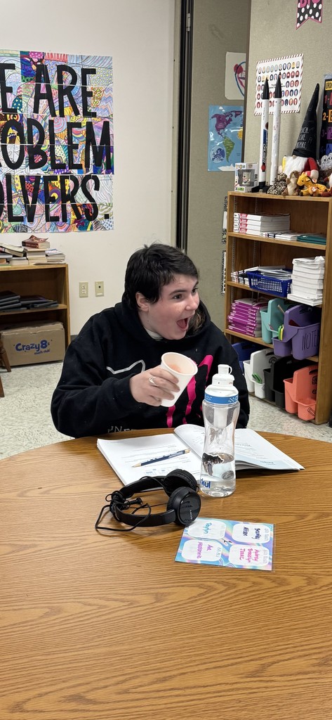  A student with dark hair and wearing a black hoodie is seated at a wooden table, looking off-camera with a wide-open, excited expression while holding a paper cup. A workbook, headphones, and a clear water bottle are on the table in front of them. A colorful poster in the background reads, "WE ARE PROBLEM SOLVERS." 