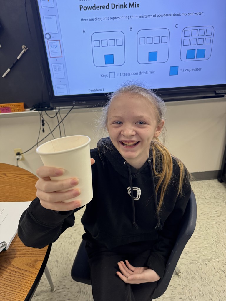 .jpg: A smiling student, a girl with her hair tied back and wearing a black hoodie, holds up a small paper cup containing a drink mixture. Behind her is a smartboard displaying a math activity tick atled "Powdered Drink Mix," which uses diagrams (A, B, C) to represent mixtures of drink mix (white squares) and water (blue squares). 