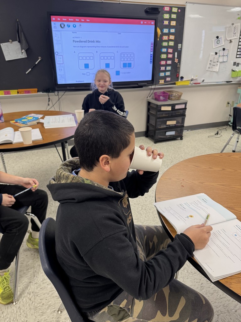  A boy in a black and camouflage jacket sits at a round table, taking a sip from a paper cup while looking down at a workbook or textbook open on the desk. In the background, another student (the girl from IMG_9209) sits near the smartboard, which displays the "Powdered Drink Mix" activity.  