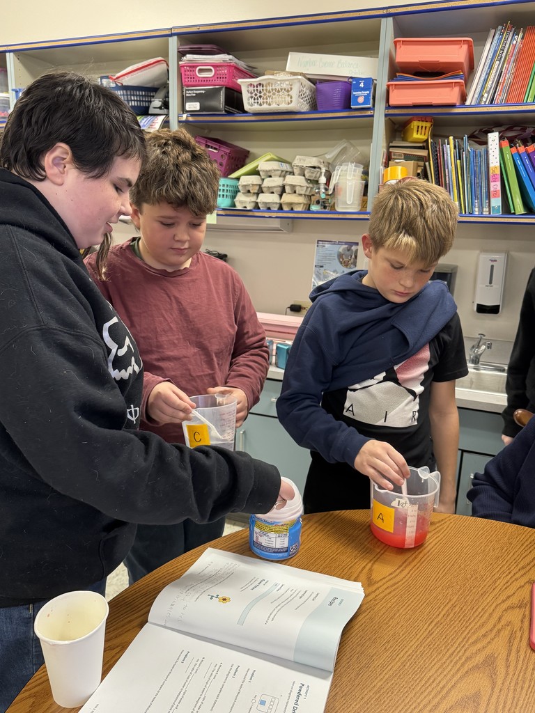  Three students stand around a desk participating in a hands-on activity. The student on the left, wearing a black hoodie, points or gestures toward a white container of powdered drink mix. The student in the middle (maroon shirt) and the student on the right (navy shirt over black shirt) are looking down and handling clear measuring cups labeled 'C' and 'A', which contain red liquid (the drink mixture). An open workbook is visible on the desk.