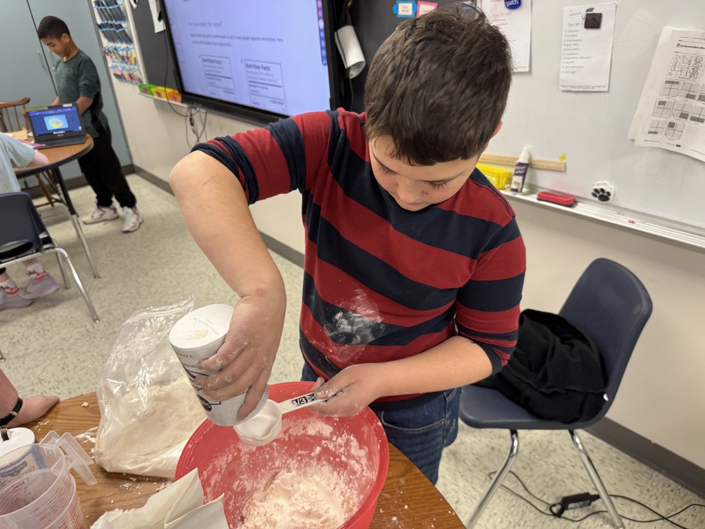 Close-up of a student pouring a white ingredient from a canister into a red mixing bowl containing flour and other ingredients, using a measuring scoop. The student is wearing a red and blue striped shirt, and the counter and shirt are dusted with flour.  