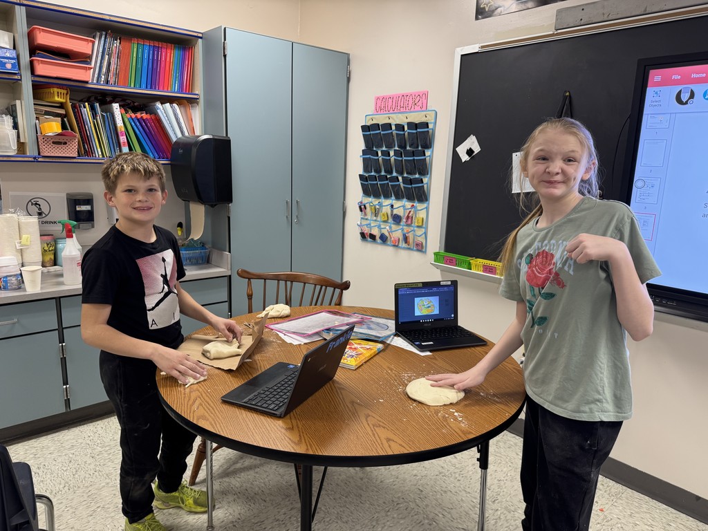 Two smiling students, a boy and a girl, stand at a round table in a classroom, each with a flattened piece of dough in front of them, seemingly for a project. They are surrounded by laptops and classroom supplies. 