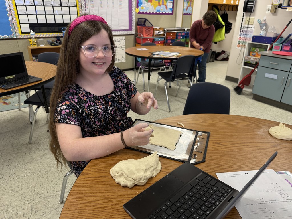 A smiling girl with long dark hair and a pink headband sits at a round table in a classroom, flattening a large piece of dough on a dark tray. A lump of dough is next to the tray, and an open laptop is also on the table.  