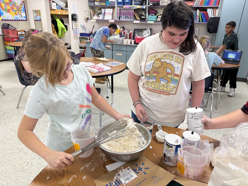 Two students working together to mix ingredients in a large silver bowl on a classroom table. The student on the left is using a whisk, while the student on the right is pouring something from a container. There are various measuring cups and ingredients on the table. 