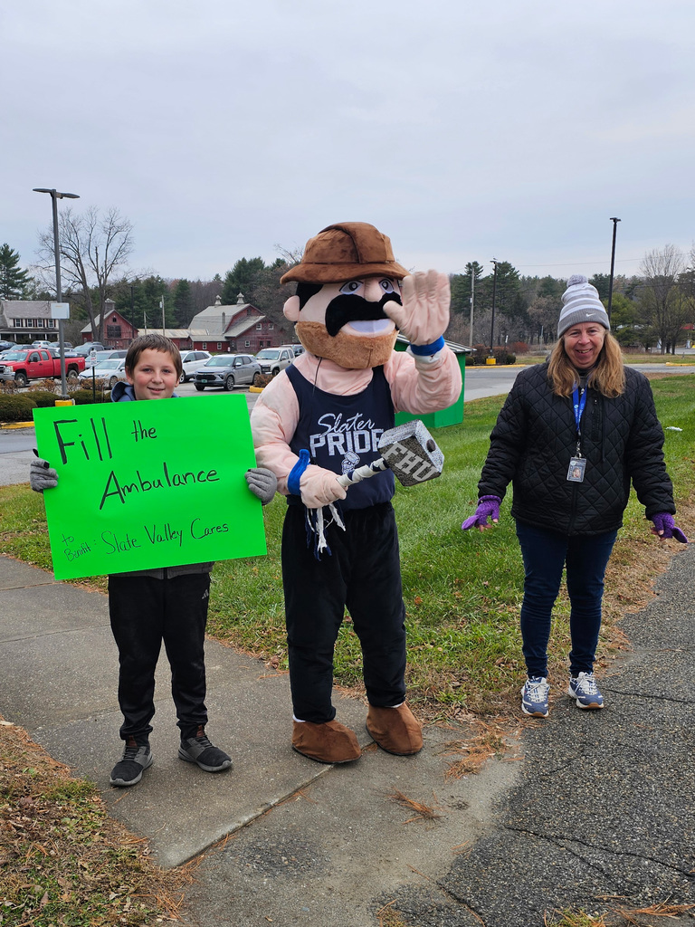 A younger student holds a bright green “Fill the Ambulance – Slate Valley Cares” sign while standing beside the Slater mascot. A staff member in a winter hat smiles on the other side. They stand near the parking lot of Fair Haven Shaw’s.