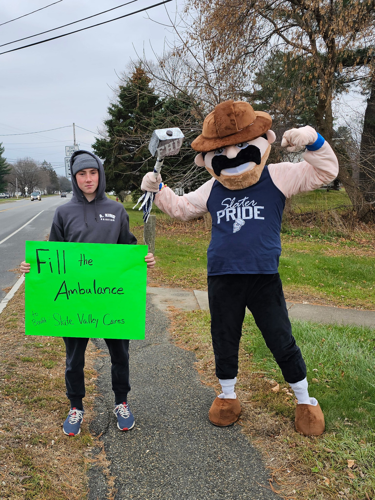 A FHUMS student stands on a sidewalk holding a bright green poster that reads “Fill the Ambulance – Slate Valley Cares.” Next to him is the Slater mascot flexing a muscle and holding a foam hammer, standing against a backdrop of a roadside and cloudy sky.
