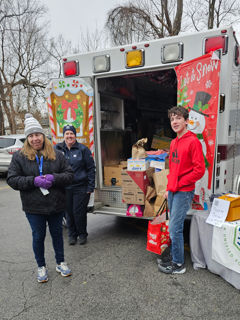 Two adults and a student stand beside the open back of a holiday-decorated ambulance filled with food and supply donations. Additional boxes and bags are arranged on the ground next to the ambulance.