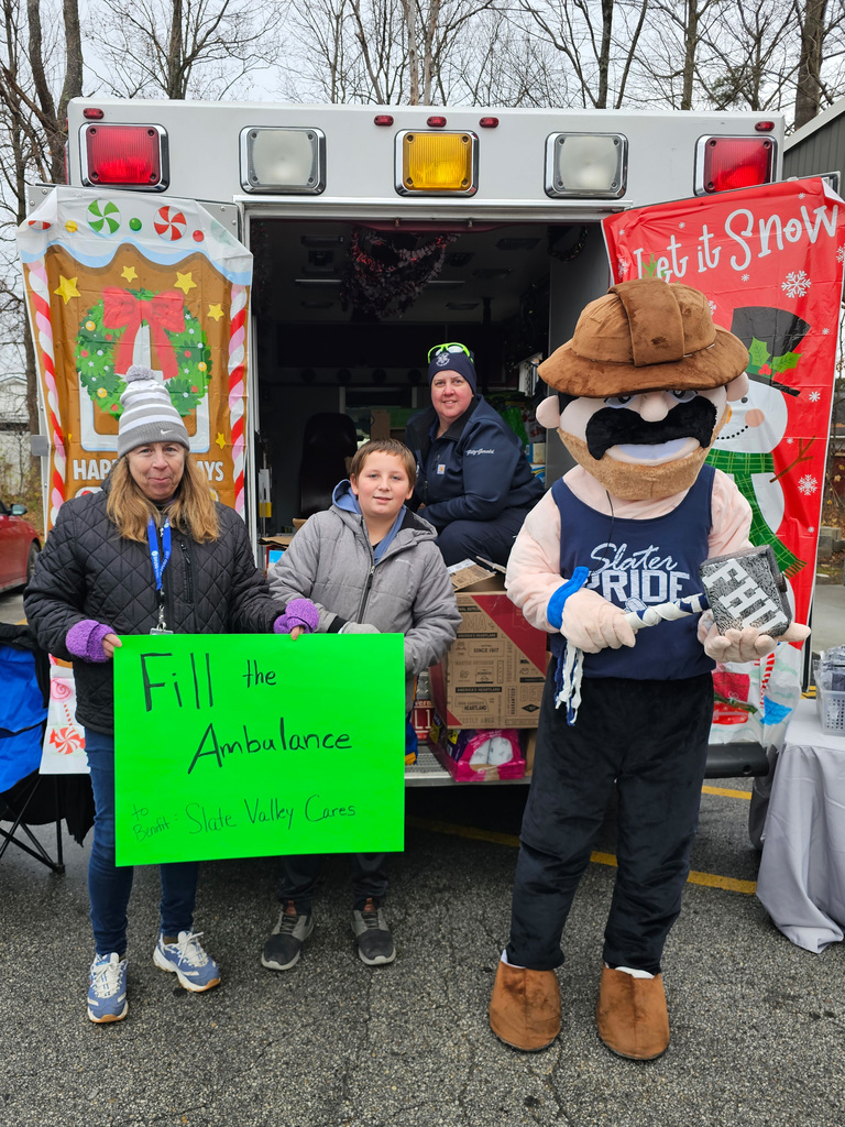 Three people and the Slater mascot stand behind an open ambulance decorated with holiday banners. They hold a bright green sign that reads “Fill the Ambulance — Slate Valley Cares.” Boxes of donated items are stacked inside the ambulance.