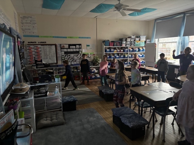 Students spread across the classroom participate in a movement activity, watching a dance routine on a TV at the front of the room.