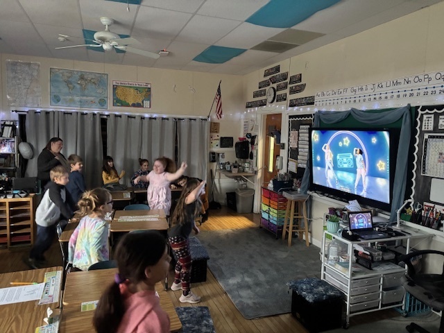 A group of elementary students in a classroom enthusiastically following a dance video on a large screen at the front of the room.
