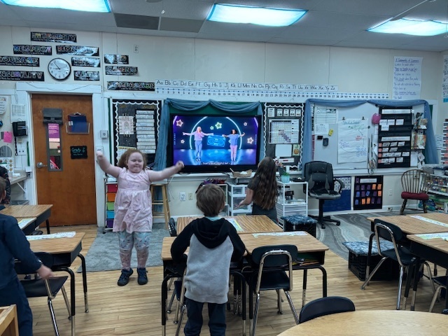 An elementary student stands smiling with arms raised while classmates follow a dance video playing on the classroom’s front screen.