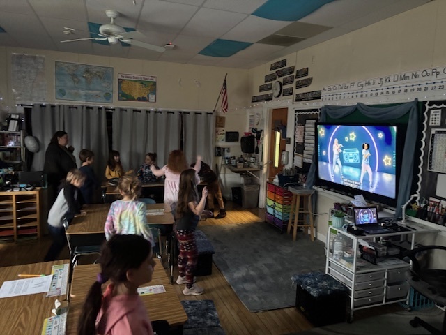 Students in a classroom follow along with a dance video on a large front screen, moving energetically in the dimly lit room.