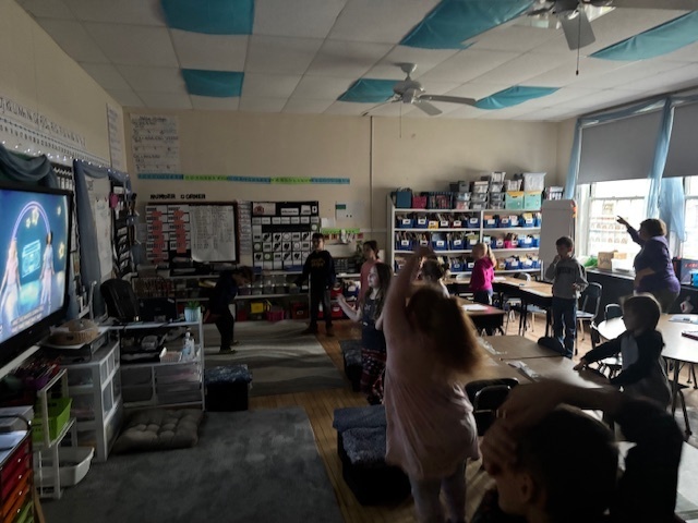 Elementary students dance along to a video displayed on a classroom screen, with several students mid-motion.