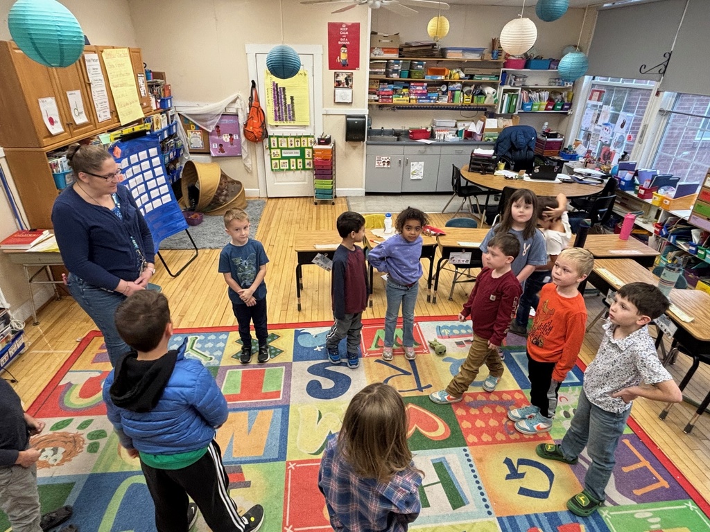 A group of young students stand in a circle on an alphabet rug as their teacher guides an activity.