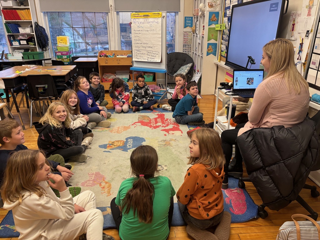 A class of students sits in a circle on a world map rug as the teacher at the front shows slides on a laptop.