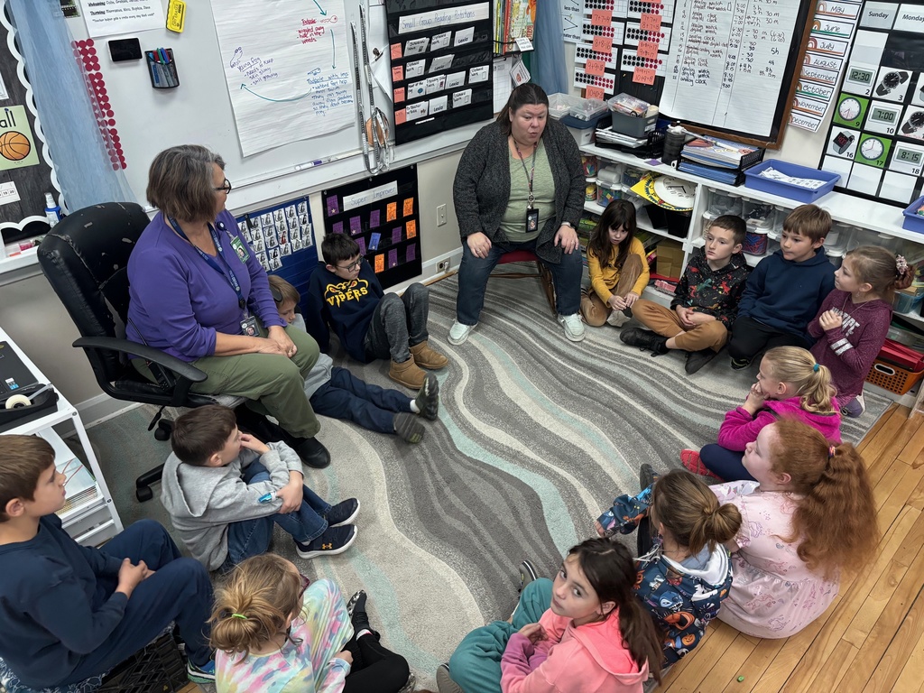 Students sit in a circle on a rug with two teachers leading a discussion at the front of the classroom.