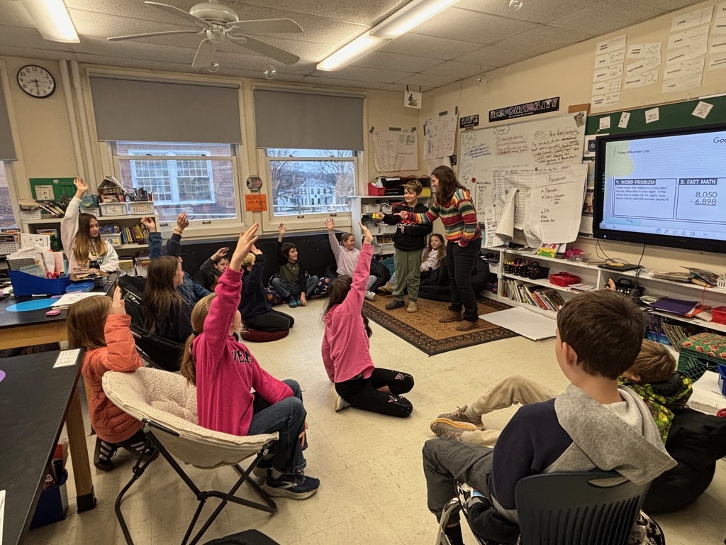 Students seated around a classroom raise their hands while a teacher at the front leads a math lesson displayed on a screen.