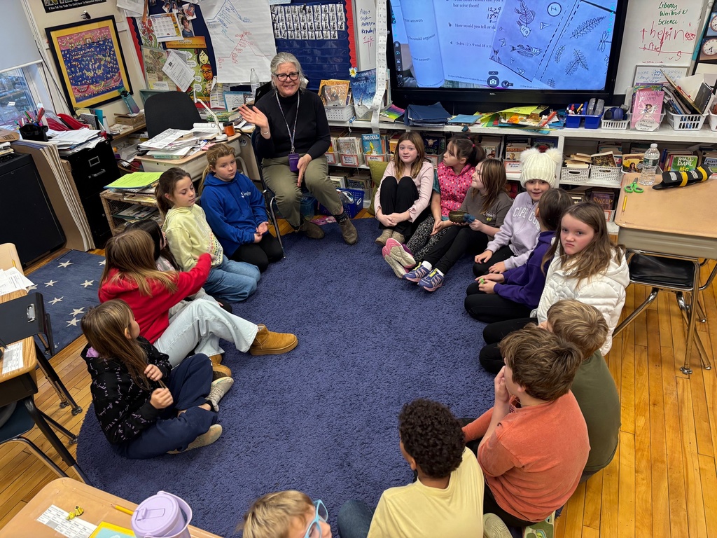 Students sit in a circle on a blue rug while a teacher leads a lesson using a large screen displaying student work.