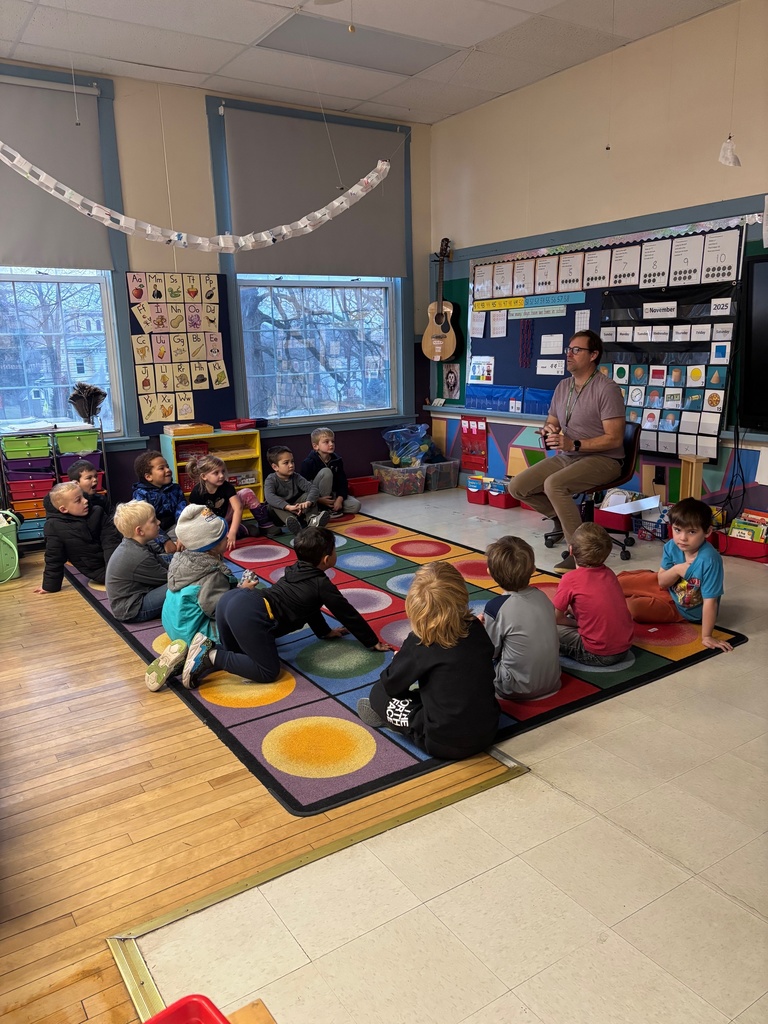 A teacher sits on a chair at the front of the room while young students sit on a colorful dot rug and listen attentively.