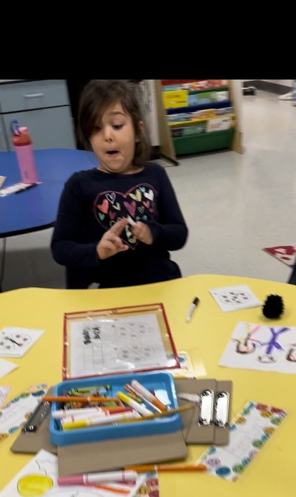  A young girl with dark hair sits at a yellow table, looking surprised with her mouth open and hands held up, holding a white marker. On the table are various papers, markers, and a tray of colored pencils. 