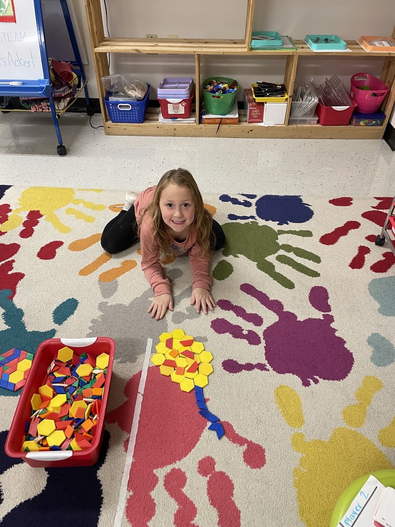 A young girl with long blonde hair lies on her stomach on a brightly colored rug patterned with large handprints, smiling at the camera. In front of her is a large arrangement of yellow and blue pattern blocks, with a red bin of additional colorful pattern blocks nearby.