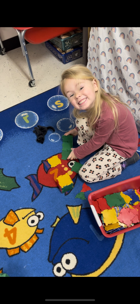  A young girl with long blonde hair smiles while sitting on a blue rug decorated with colorful fish. She is playing with green and yellow interlocking square blocks, with a red bin full of colorful blocks next to her.