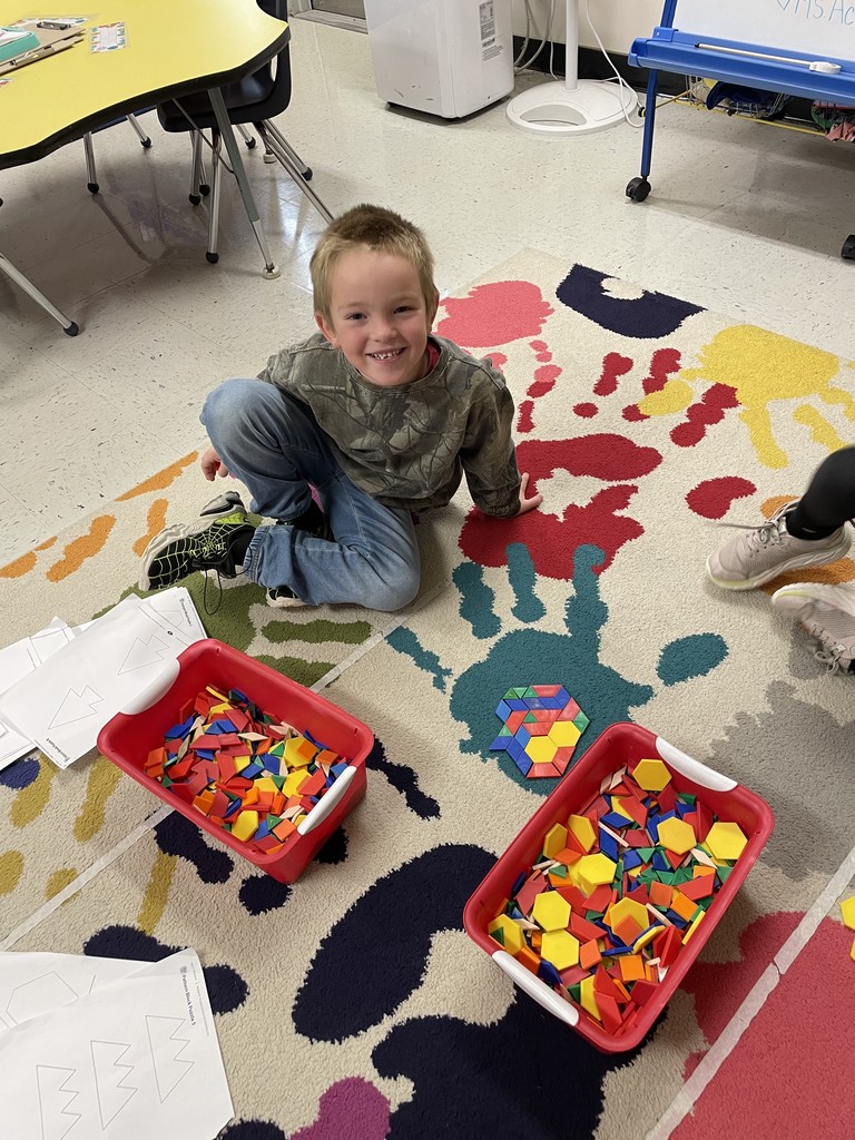  A young boy wearing a camouflage shirt and jeans sits on a brightly colored rug patterned with large handprints, smiling at the camera. In front of him are two red bins filled with colorful pattern blocks, with a partially completed geometric design visible on the rug.  