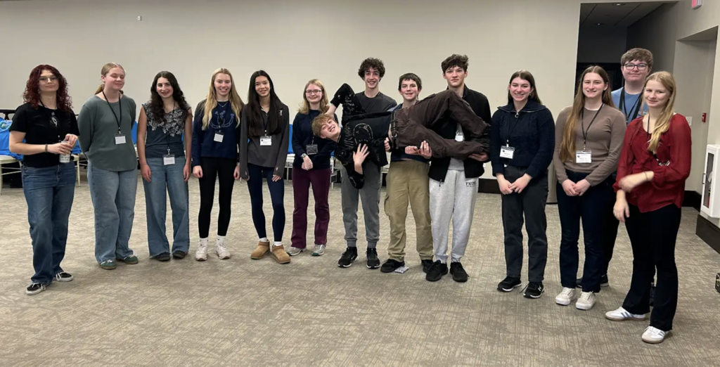 High school students standing in a row inside a large room, smiling; one student is being held horizontally by two classmates in a playful pose.