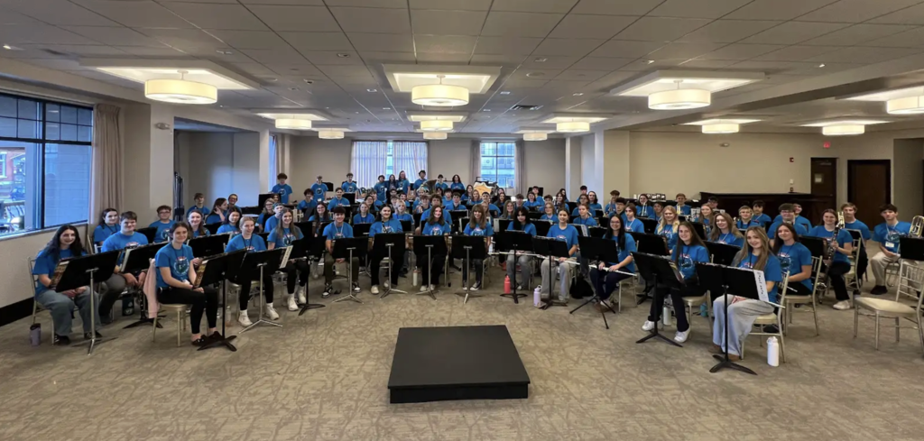 A large group of high school musicians seated with music stands in a spacious rehearsal room, all wearing matching blue T-shirts and facing the conductor’s podium.