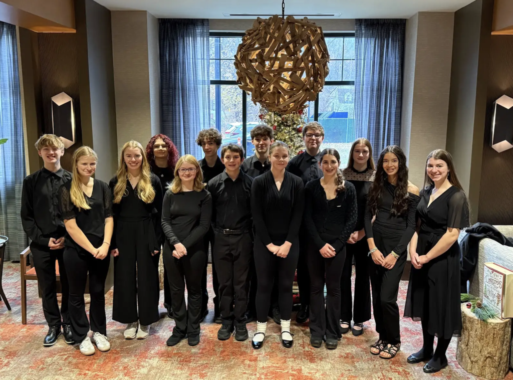 Group photo of high school musicians dressed in black concert attire, standing together in a hotel lobby decorated with a holiday tree and large hanging wooden light fixture.