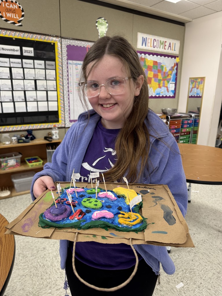 A smiling girl holding a colorful, 3D model of a cell made from clay or dough on a brown paper bag base." 