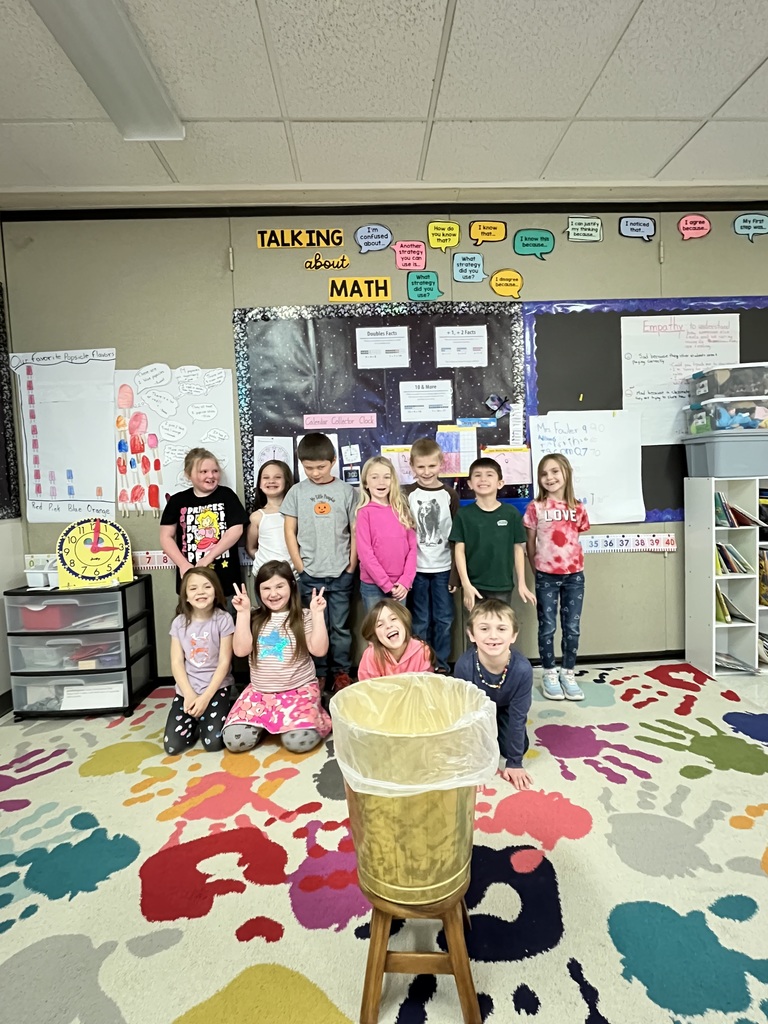 A group of elementary school children, some kneeling and some standing, smile at the camera in a colorful classroom. In the foreground, a tall, golden trash can with a plastic liner sits on a wooden stool. The floor is covered with a whimsical rug featuring irregular splatters of red, blue, green, and orange. On the left, five girls are arranged: two kneeling in front and three standing behind. The girl on the far left wears a black shirt with colorful graphics, while next to her, a girl in a light purple shirt makes a peace sign. Another girl in a pink and white tie-dye shirt kneels with a peace sign, and beside her, a girl in a pink long-sleeved shirt laughs with her hands near her face. Behind them, a girl in a white top smiles. On the right side of the photo, four boys stand in a row. From left to right, they wear a grey t-shirt, a white t-shirt with graphics, a dark green polo shirt, and a blue long-sleeved shirt. The classroom walls are adorned with various educational posters. A large black bulletin board in the center has the title "TALKING about MATH" at the top and features charts and papers, some with a space theme. To the right of the bulletin board, another board displays colorful shapes and text. On the far left, a white board shows illustrations of what appear to be organs. A yellow clock and a stack of colorful bins are visible on a small table on the left.