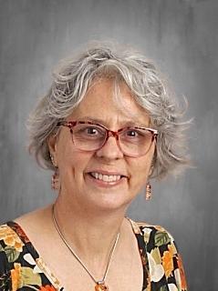 A portrait of a woman with short curly gray hair and red-framed glasses, smiling at the camera. She is wearing a floral top with orange and yellow flowers and small drop earrings, posed in front of a gray studio background.