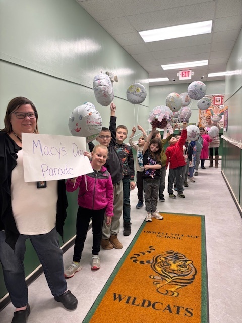 A group of elementary students stands in a school hallway holding handmade Thanksgiving-themed balloons for a “Macy’s Day Parade” activity. A teacher in front holds a sign labeled “Macy’s Day Parade.” The students smile as they line up along a floor mat featuring a wildcat mascot.