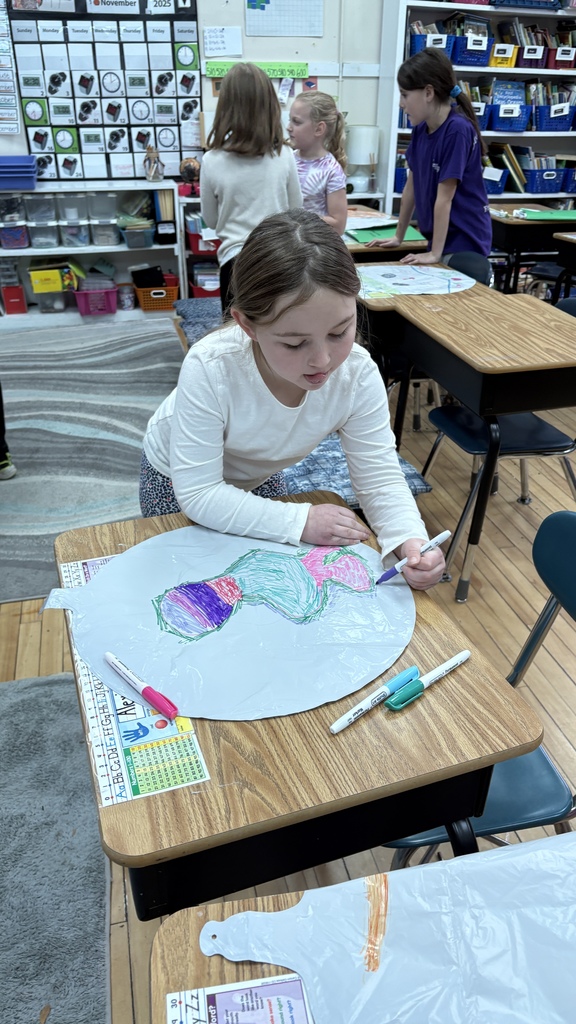 A young student sits at her desk coloring a large balloon-shaped plastic cutout with bright markers. Other students work in the background inside a classroom filled with learning materials and organized bins.