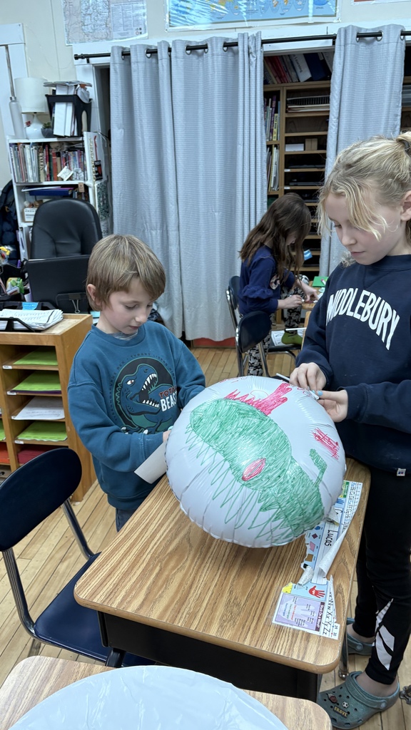 Two students work together at a desk, coloring a large balloon-shaped plastic cutout. One student holds the balloon while the other colors it with green and red marker. The classroom shelves and materials are visible behind them.
