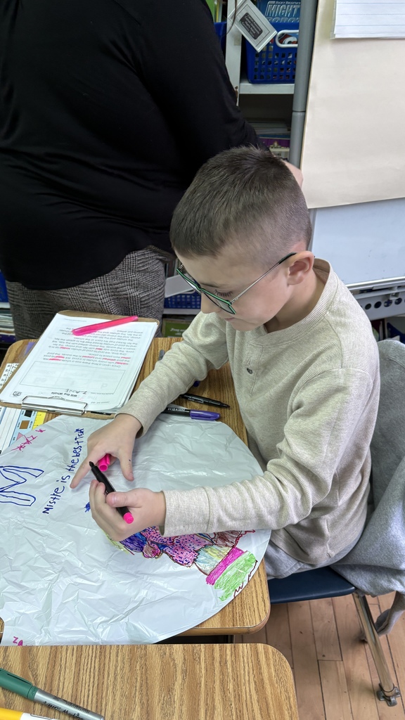A student wearing glasses colors a balloon-shaped plastic cutout with brightly colored markers. He is focused on his artwork, with worksheets, markers, and classroom supplies spread out on the desk around him.