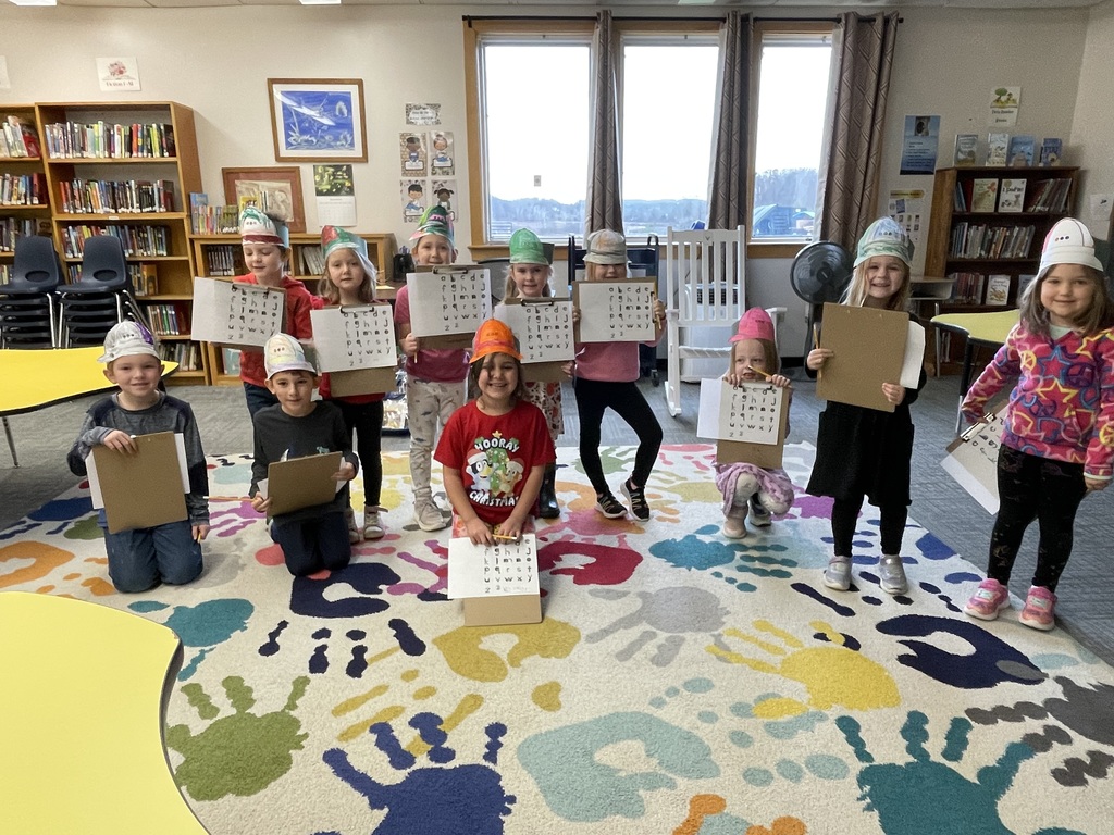 A group of approximately ten young children, all wearing festive paper hats (some green, some light blue/white), pose for a photo in a library. They are holding clipboards with papers that have letters and symbols visible.