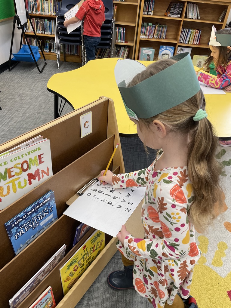 A young girl in a white dress with a pumpkin and leaf print stands next to a wooden bookshelf, using the shelf as a surface to write on a paper attached to a clipboard. She is wearing a green paper headband.