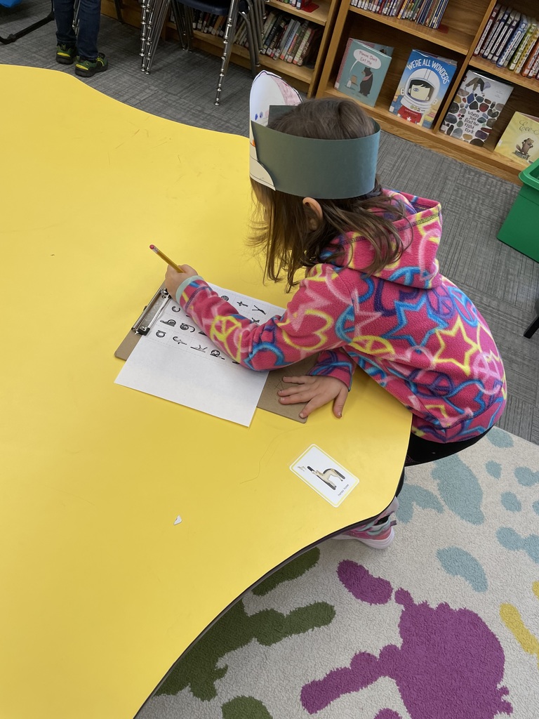 A young girl in a bright pink and blue star-patterned fleece jacket is seated at a yellow table, concentrating as she writes on a paper attached to a clipboard. A card with a letter 'H' is on the table next to her. IMG_4989.jpg: A young girl in a red shirt and a decorated paper hat stands next to a tall bookshelf, holding a pencil and pointing at a small card with a picture on it. She has a clipboard with a paper that has letters and symbols.