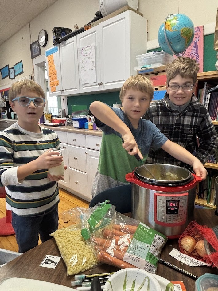 Three boys stand around an Instant Pot full of soup, with one stirring it. Various ingredients—including carrots, onions, and pasta—are spread across the classroom table.