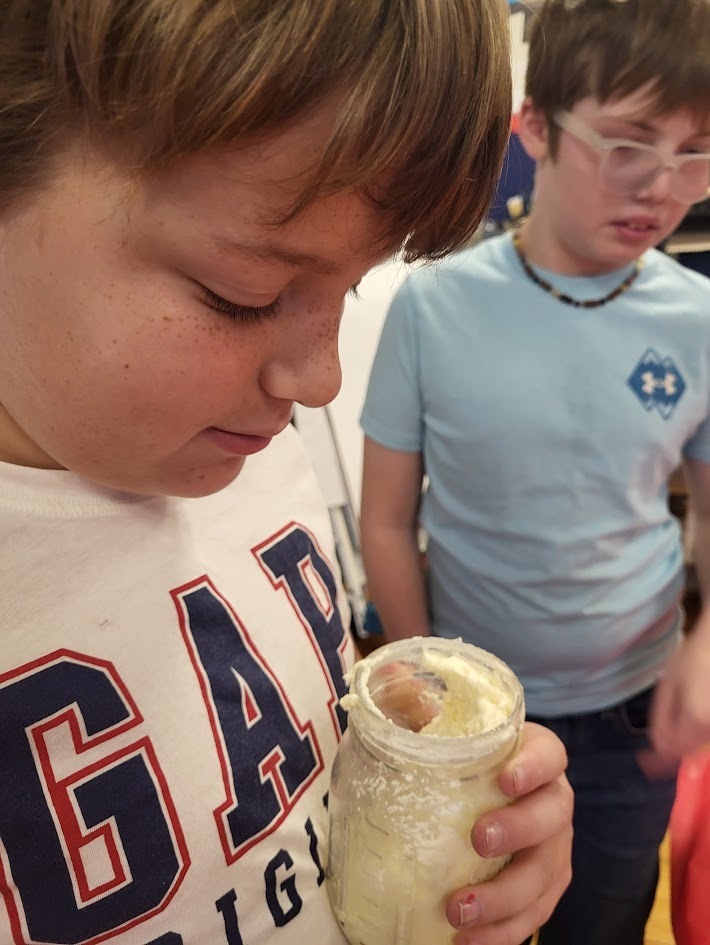 A close-up of a student examining a jar of freshly made butter. Another child stands blurred in the background.