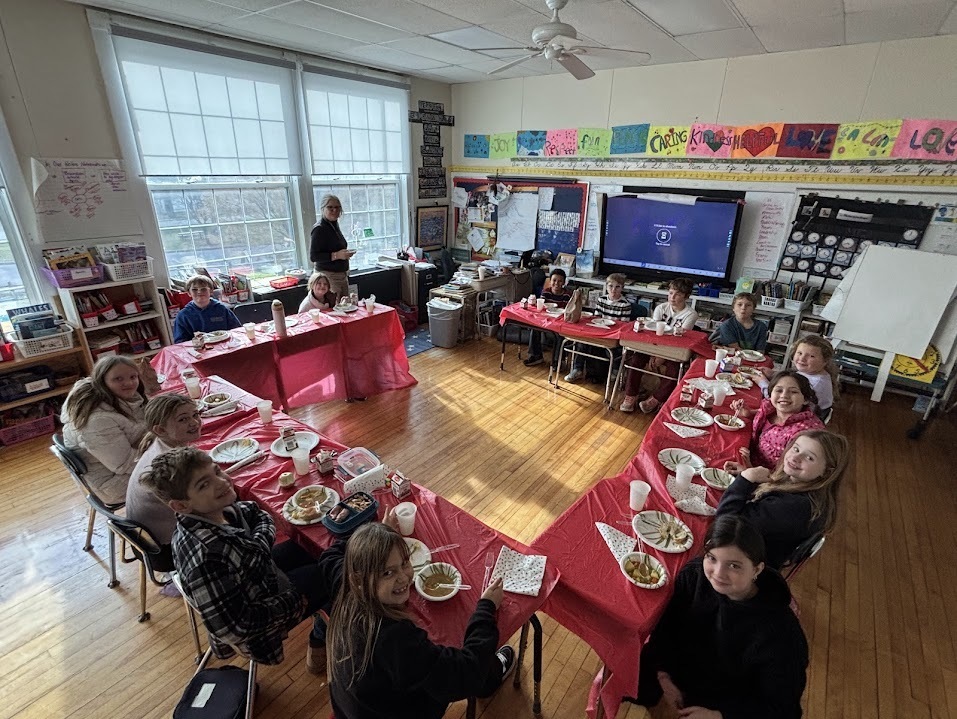 A classroom of students sits around tables arranged in a square, eating a meal served in bowls and cups. A teacher stands near the back wall beside a large screen.