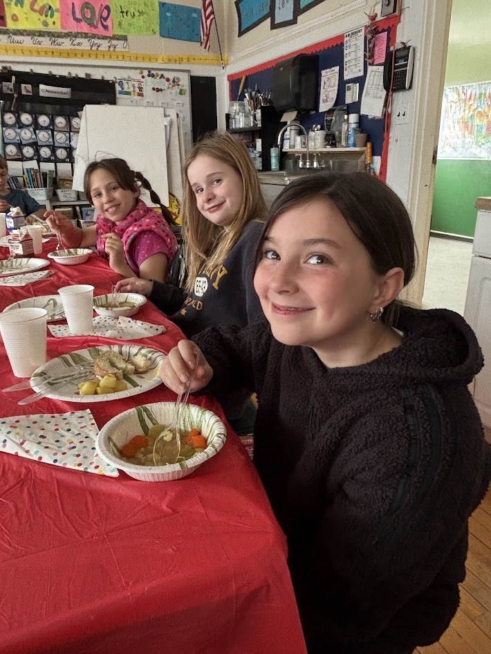 Three girls smile while eating soup at a long table covered in red tablecloths. Plates, cups, and polka-dot napkins are arranged in front of them.