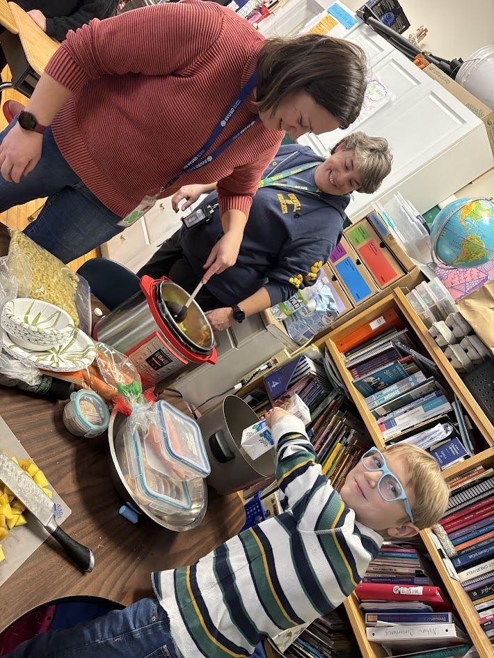 Two adults assist a young boy with glasses as he pours ingredients into a pot on a classroom table filled with vegetables, containers, and cooking supplies. Bookshelves and classroom materials surround them.
