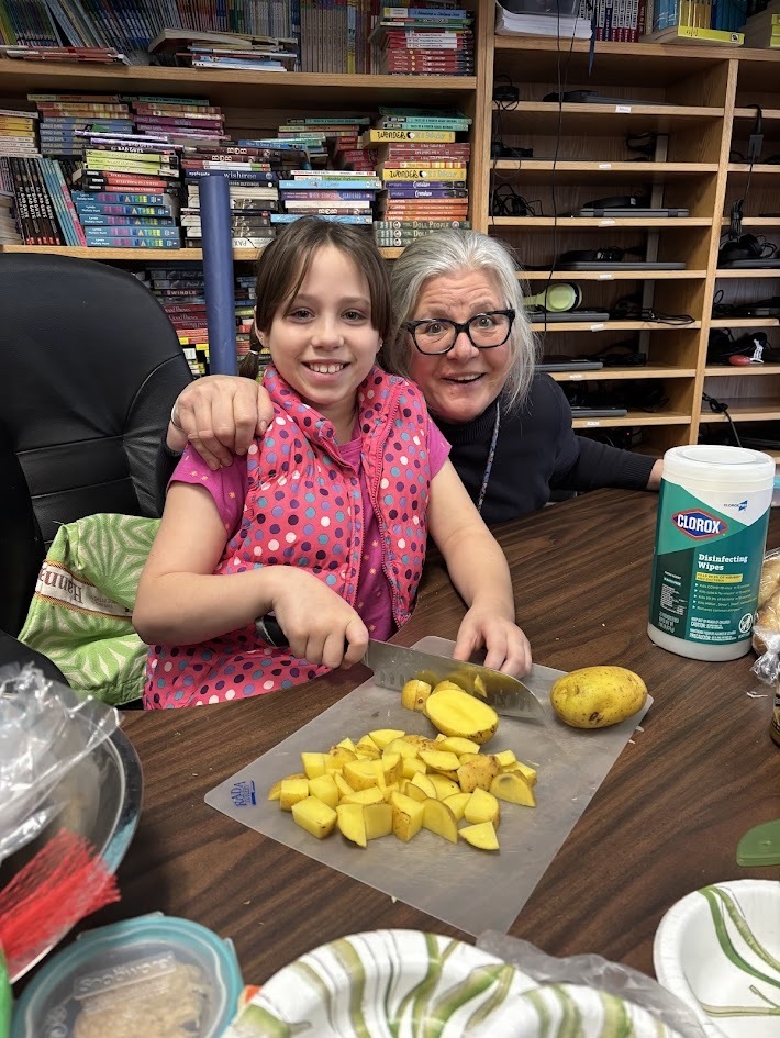 A young student in a pink patterned vest smiles while chopping yellow potatoes at a table. An adult woman with gray hair and glasses hugs her from behind, both smiling. Bookshelves filled with colorful books line the background.