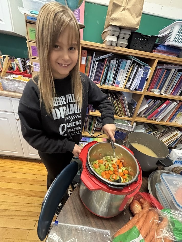 A smiling young girl in a black sweatshirt stirs chopped vegetables—including carrots and celery—in an Instant Pot on a classroom table filled with cooking supplies and vegetables.